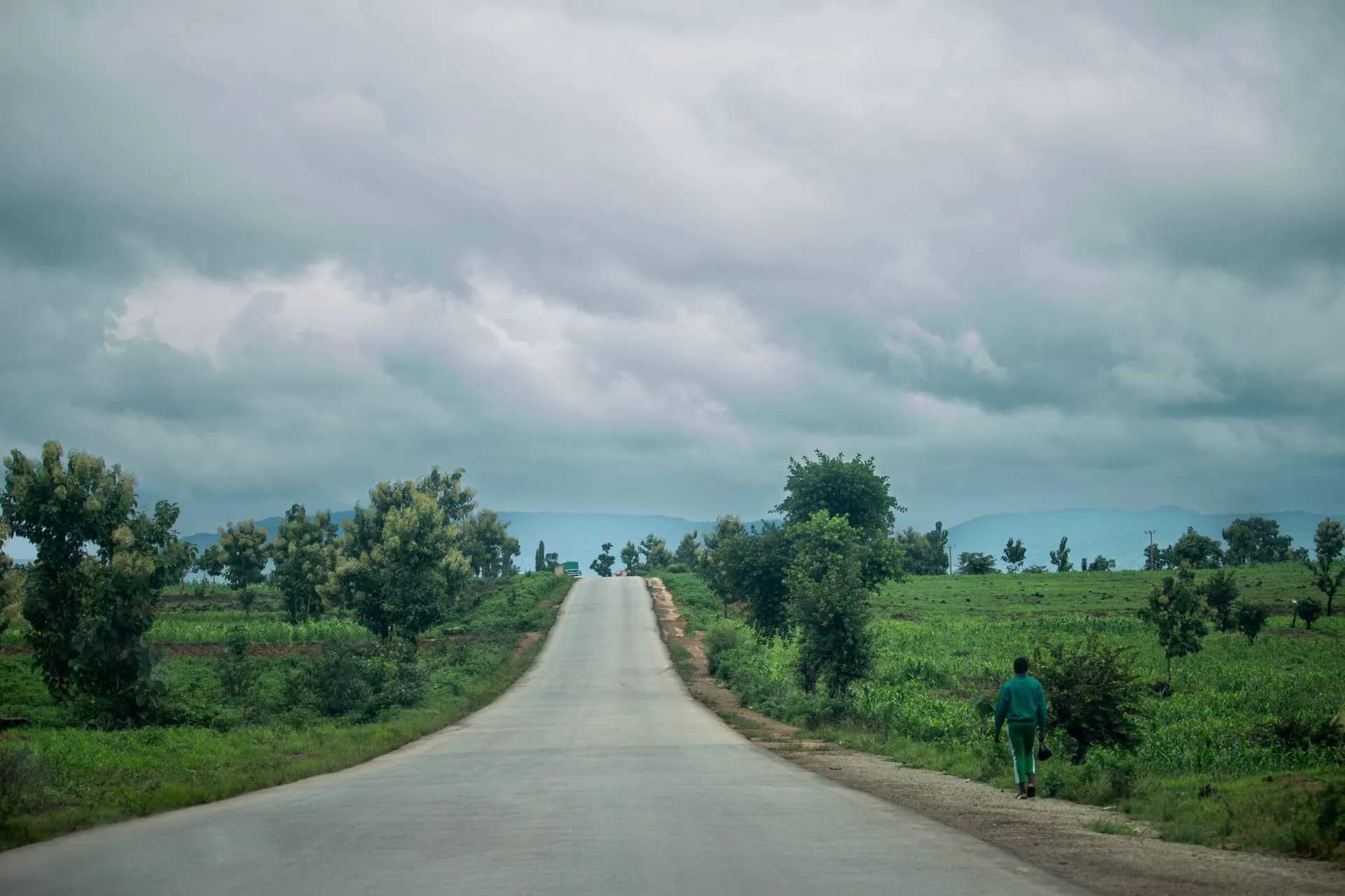 An open road running through green farmland under a wide, clouded sky, one person walking its shoulder