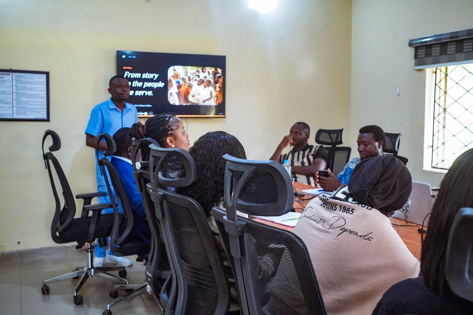 A facilitator presents to a team seated around a table with laptops, a screen behind him.