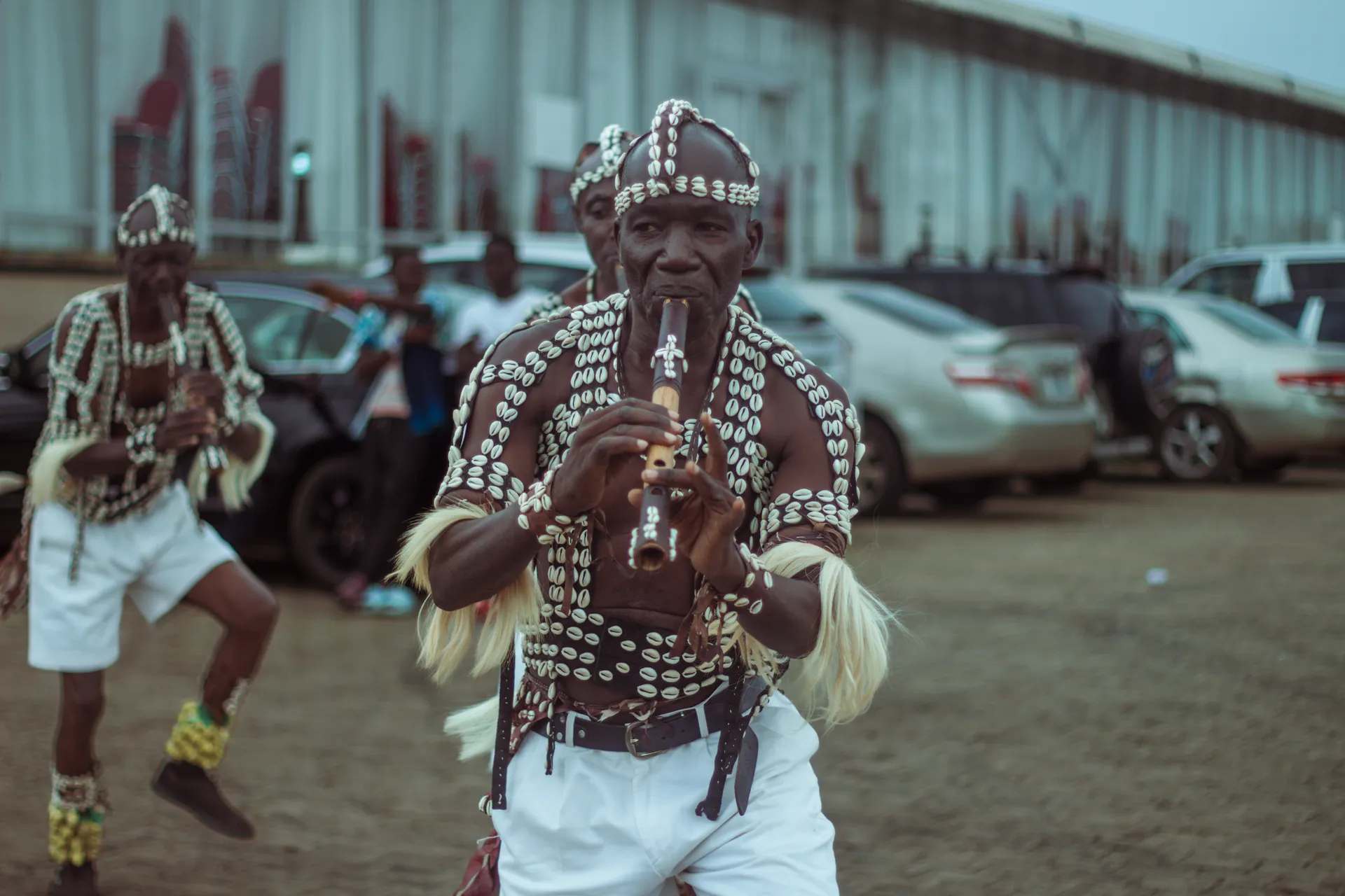 A performer in beaded regalia and a cowrie headdress playing a flute, parked cars behind him