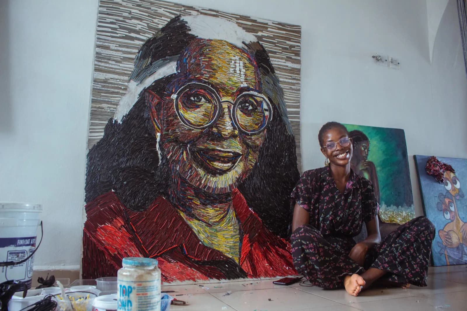 A smiling artist sits in her studio beside a large painted portrait.